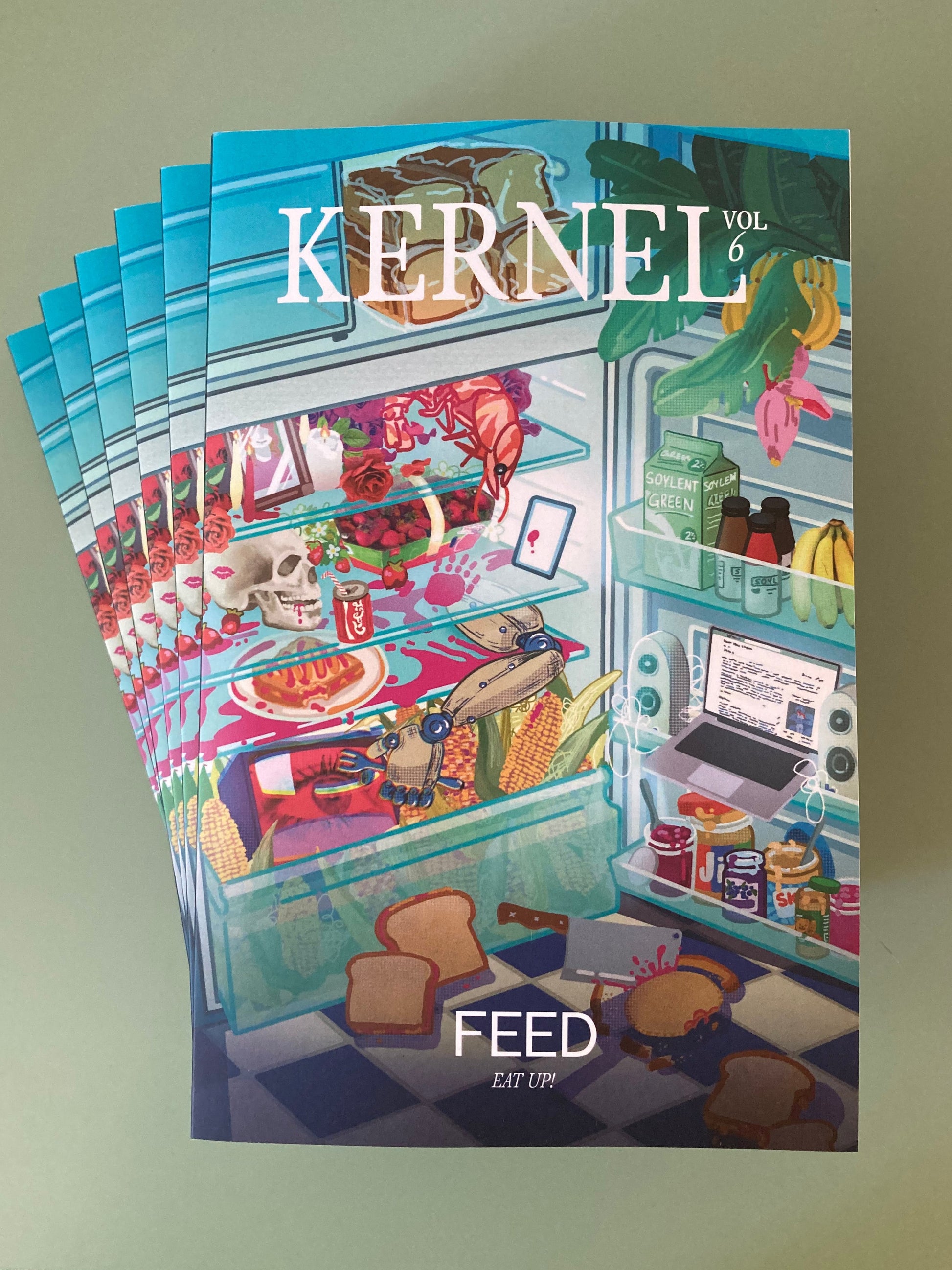 A fanned stack of KERNEL 6: FEED copies featuring a colorful, surreal illustration by Kristy Gao of a kitchen scene with food items, displayed on a light green surface.